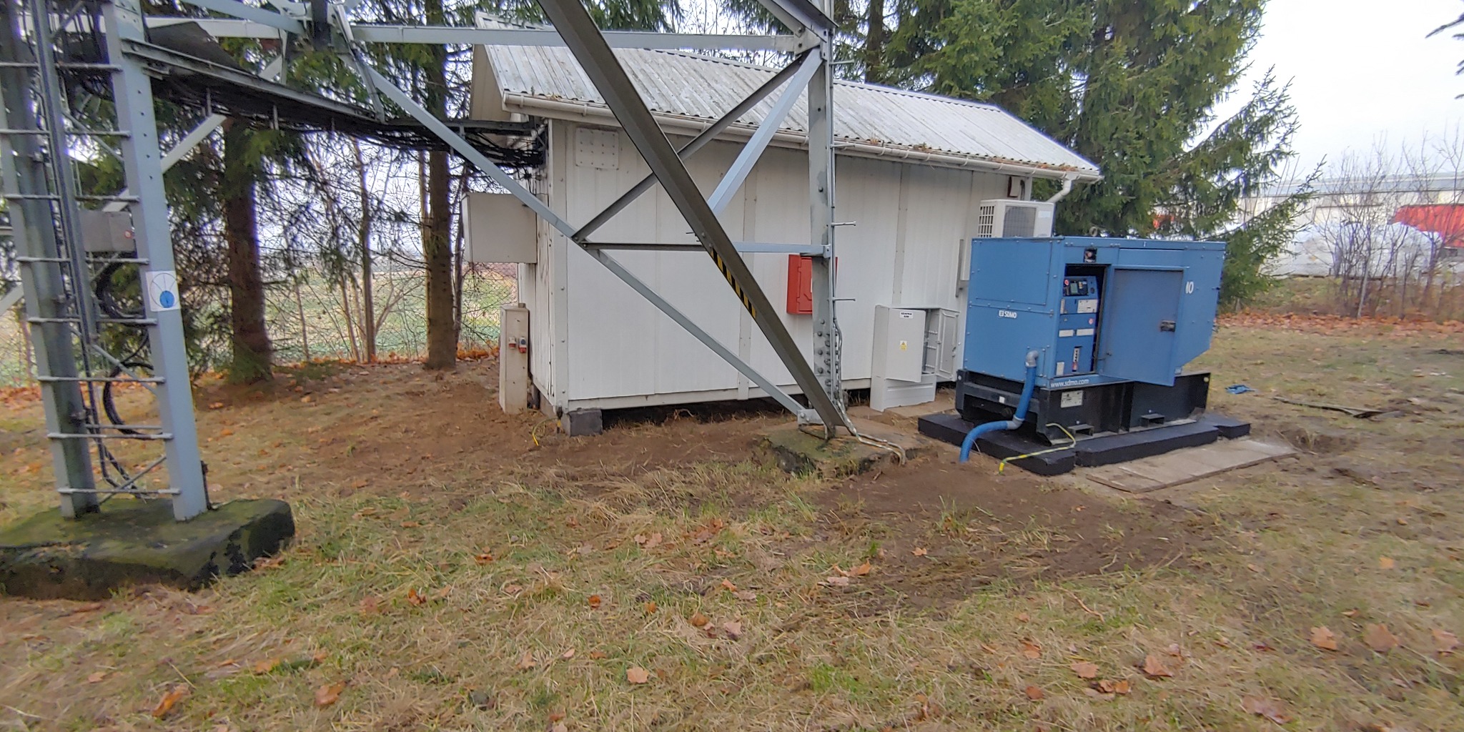 Agregat prądotwórczy obok stacji transformatorowej z metalową konstrukcją wsporczą, widoczne elementy instalacji elektrycznej i przyłącza.