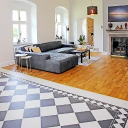 Przestronny salon z szarym narożnikiem, kominkiem z dekoracjami, drewnianą podłogą i czarno-białą szachownicą z płytek przy ścianie.