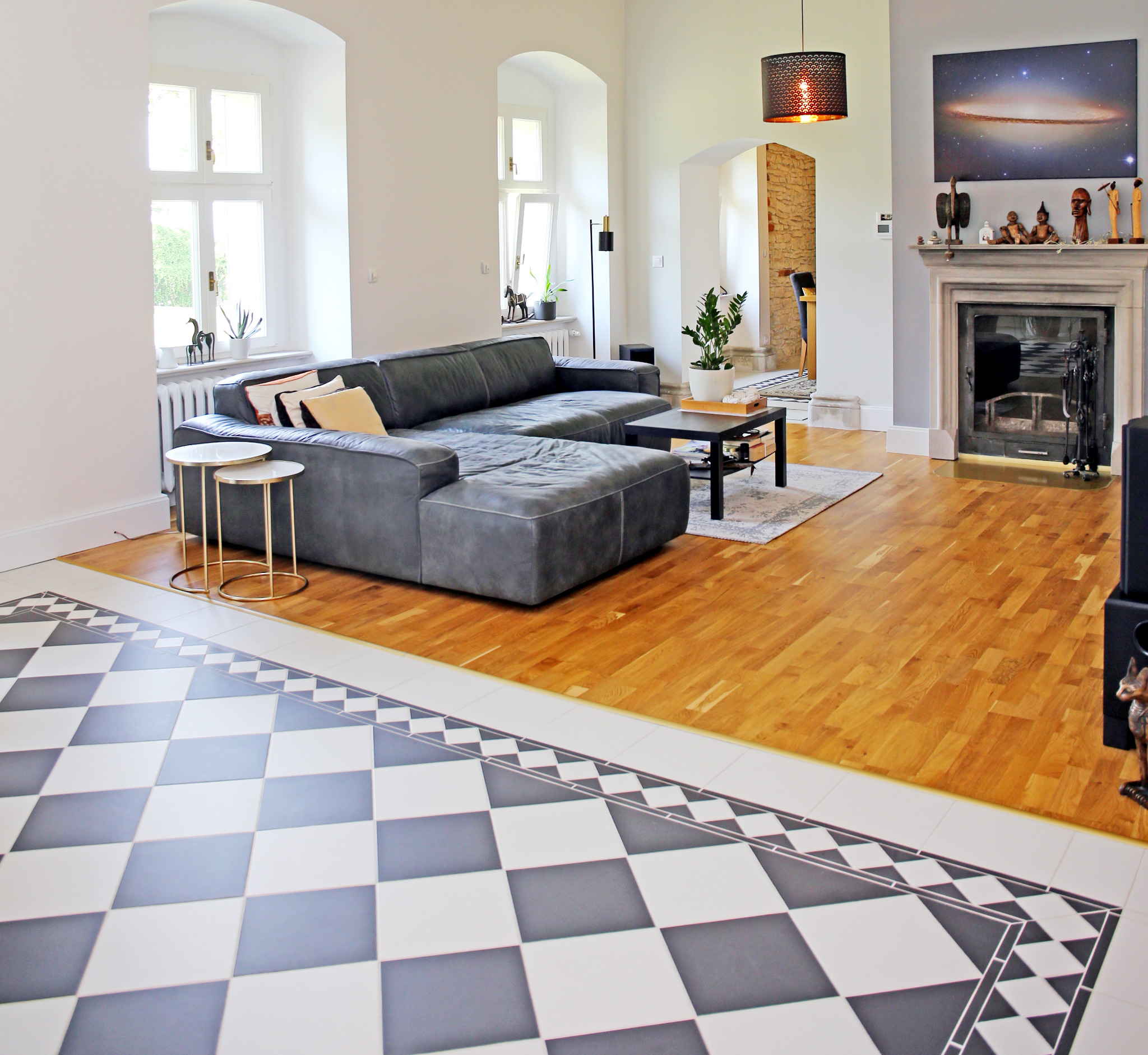 Przestronny salon z szarym narożnikiem, kominkiem z dekoracjami, drewnianą podłogą i czarno-białą szachownicą z płytek przy ścianie.