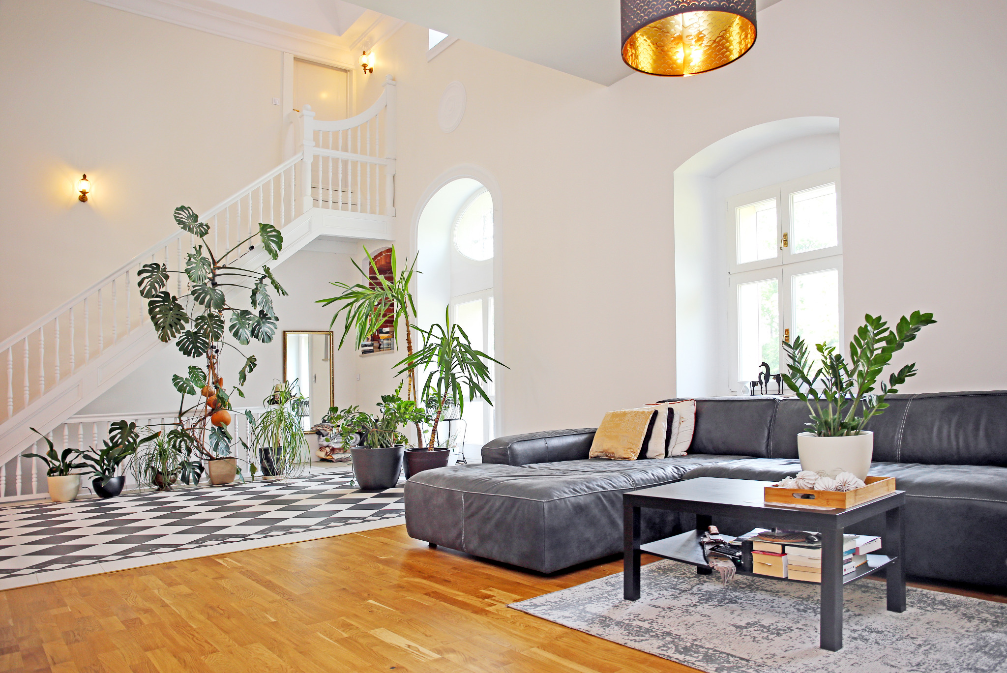 Jasne, przestronne wnętrze z wysokim sufitem, białymi ścianami, czarno-białą podłogą w szachownicę, drewnianą podłogą, schodami z białymi balustradami, szarą skórzaną sofą i licznymi roślinami...