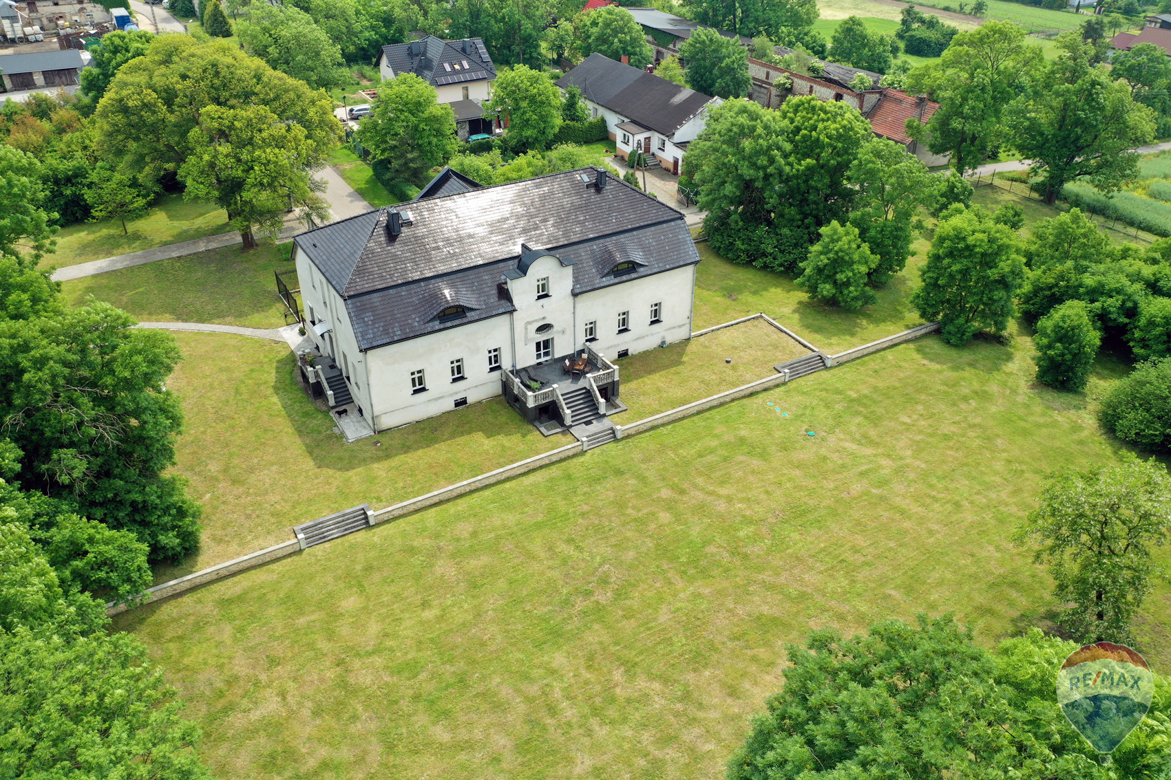 Zdjęcie z drona przedstawia duży, biały, dwupiętrowy dom z ciemnym dachem, otoczony rozległym, zielonym trawnikiem i drzewami. Widoczny mur oporowy z kamienia. Na trawniku widoczne ślady po zabawie...