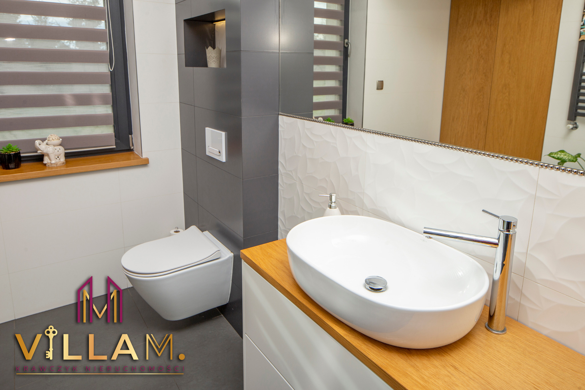 Nowoczesna łazienka z owalną umywalką nablatową, drewnianym blatem i minimalistyczną toaletą w zabudowie, uzupełniona dekoracyjnymi figurkami na parapecie okna.