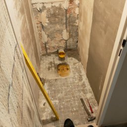 Łukasz Zduńczyk - Łazienka w trakcie remontu: widoczne stare płytki podłogowe, częściowo skuwa ściana z odsłoniętą cegłą, żółty rura kanalizacyjna, poziomica oparta o ścianę, młotek i śrubokręt na podłodze.