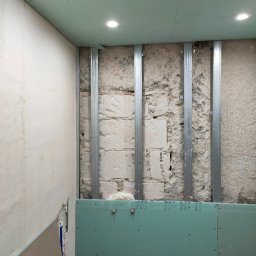 Łukasz Zduńczyk - Wnętrze w trakcie prac: montaż stelaża i płyt gipsowo-kartonowych na ścianach i suficie, widoczne profile stalowe i fragmenty odsłoniętej, surowej ściany z cegieł.