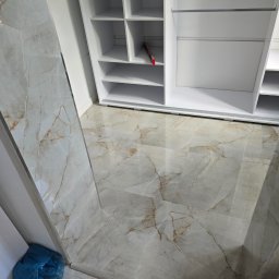 Łukasz Zduńczyk - Ułożona podłoga z płytek imitujących marmur w odcieniach szarości i beżu, częściowo widoczna biała szafa wnękowa z półkami, w rogu niebieski worek na śmieci.