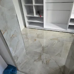 Ułożona podłoga z płytek imitujących marmur w odcieniach szarości i beżu, częściowo widoczna biała szafa wnękowa z półkami, w rogu niebieski worek na śmieci.