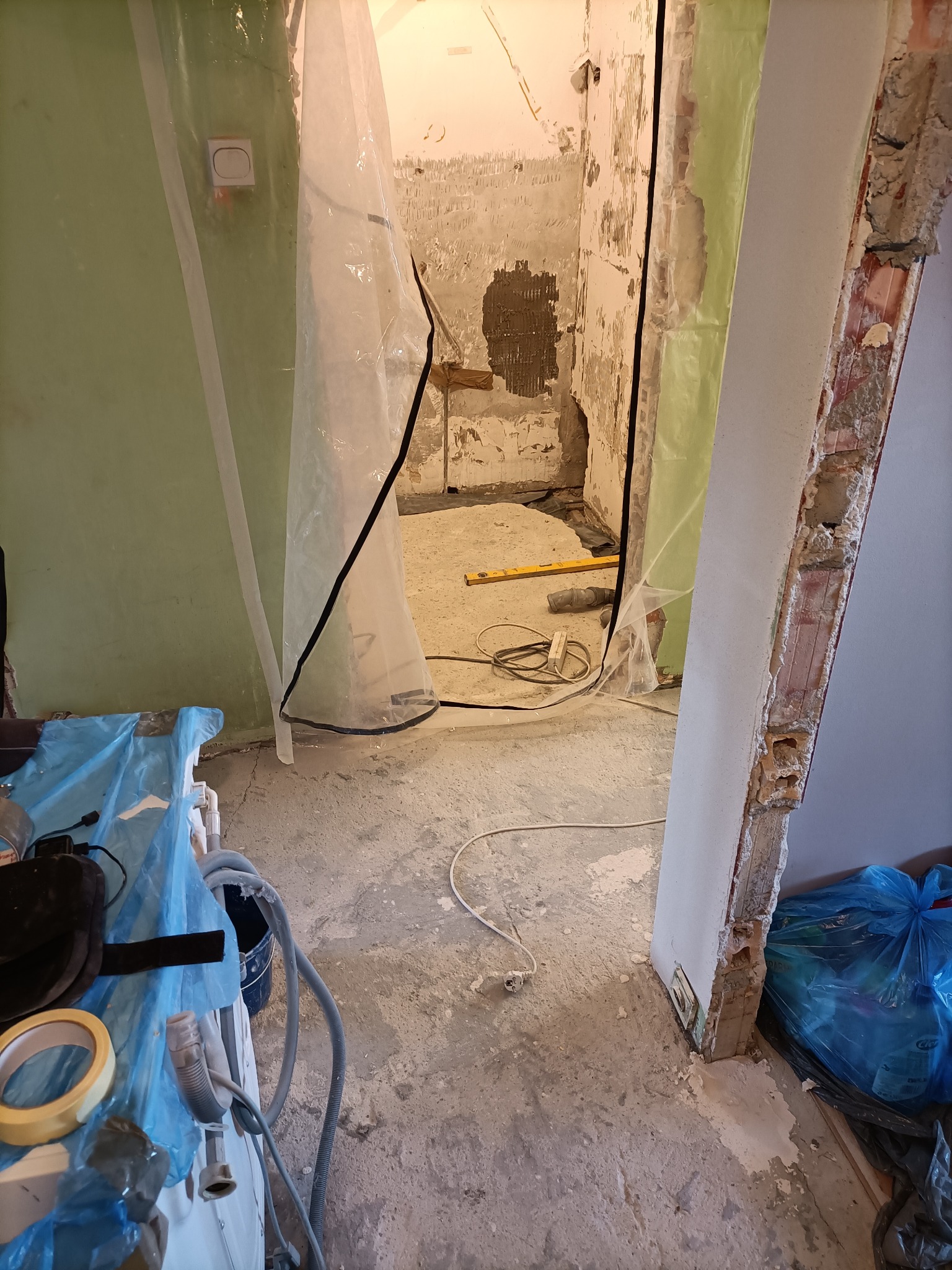 Łazienka w trakcie demontażu: zerwane płytki, zabezpieczone folią drzwi i ściany, widoczne narzędzia i gruz.