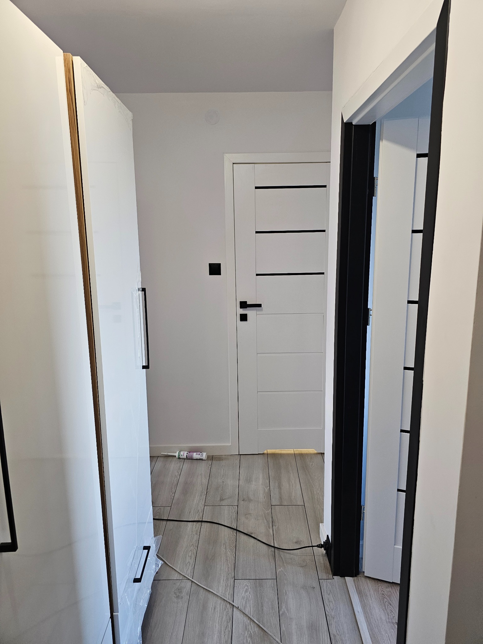 Wnętrze mieszkania w trakcie wykańczania: widoczna biała szafa z czarnymi uchwytami w folii ochronnej, białe drzwi z czarnymi poziomymi paskami, czarna ościeżnica, szare panele podłogowe oraz tuba...