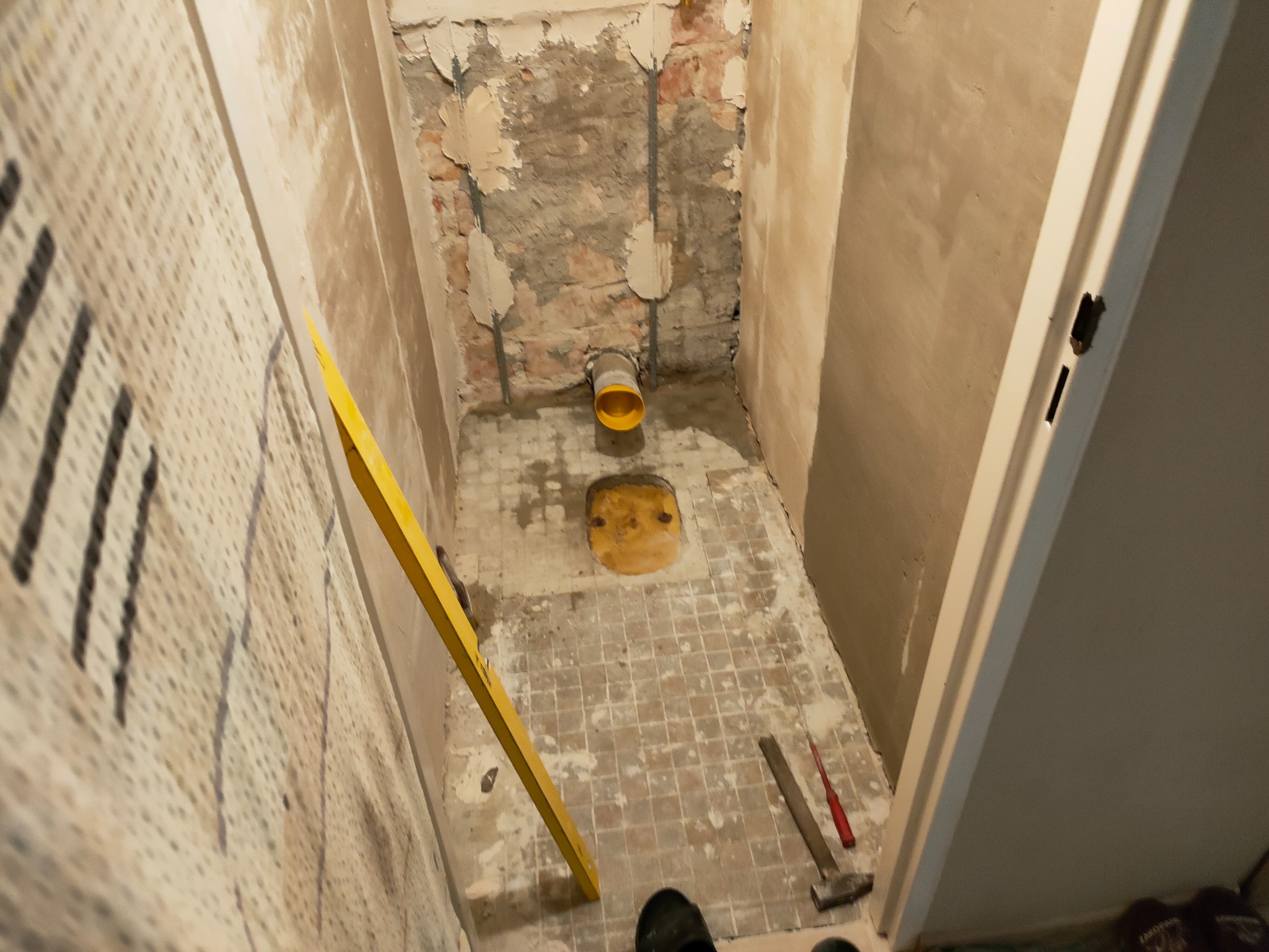 Łazienka w trakcie remontu: widoczne stare płytki podłogowe, częściowo skuwa ściana z odsłoniętą cegłą, żółty rura kanalizacyjna, poziomica oparta o ścianę, młotek i śrubokręt na podłodze.