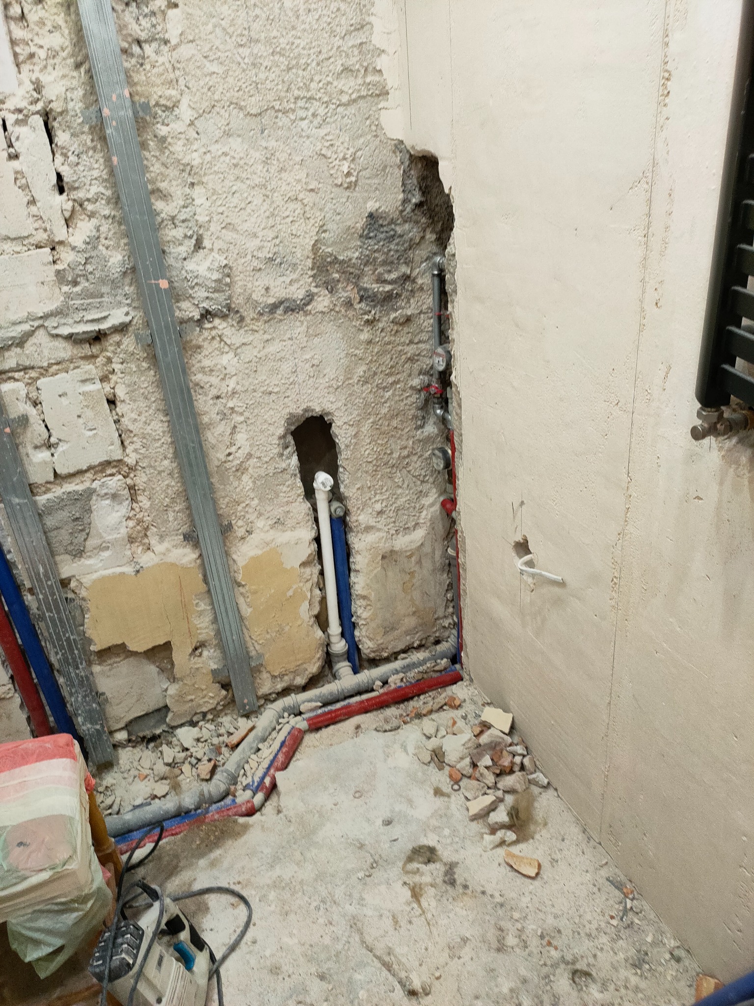 Fragment ściany w trakcie prac instalacyjnych w łazience, widoczne rury wodne (czerwone i niebieskie), metalowe profile do montażu płyt gipsowo-kartonowych oraz gruz na podłodze.