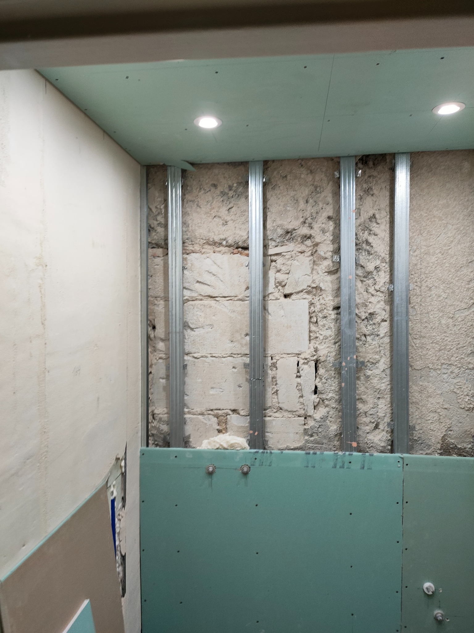 Wnętrze w trakcie prac: montaż stelaża i płyt gipsowo-kartonowych na ścianach i suficie, widoczne profile stalowe i fragmenty odsłoniętej, surowej ściany z cegieł.