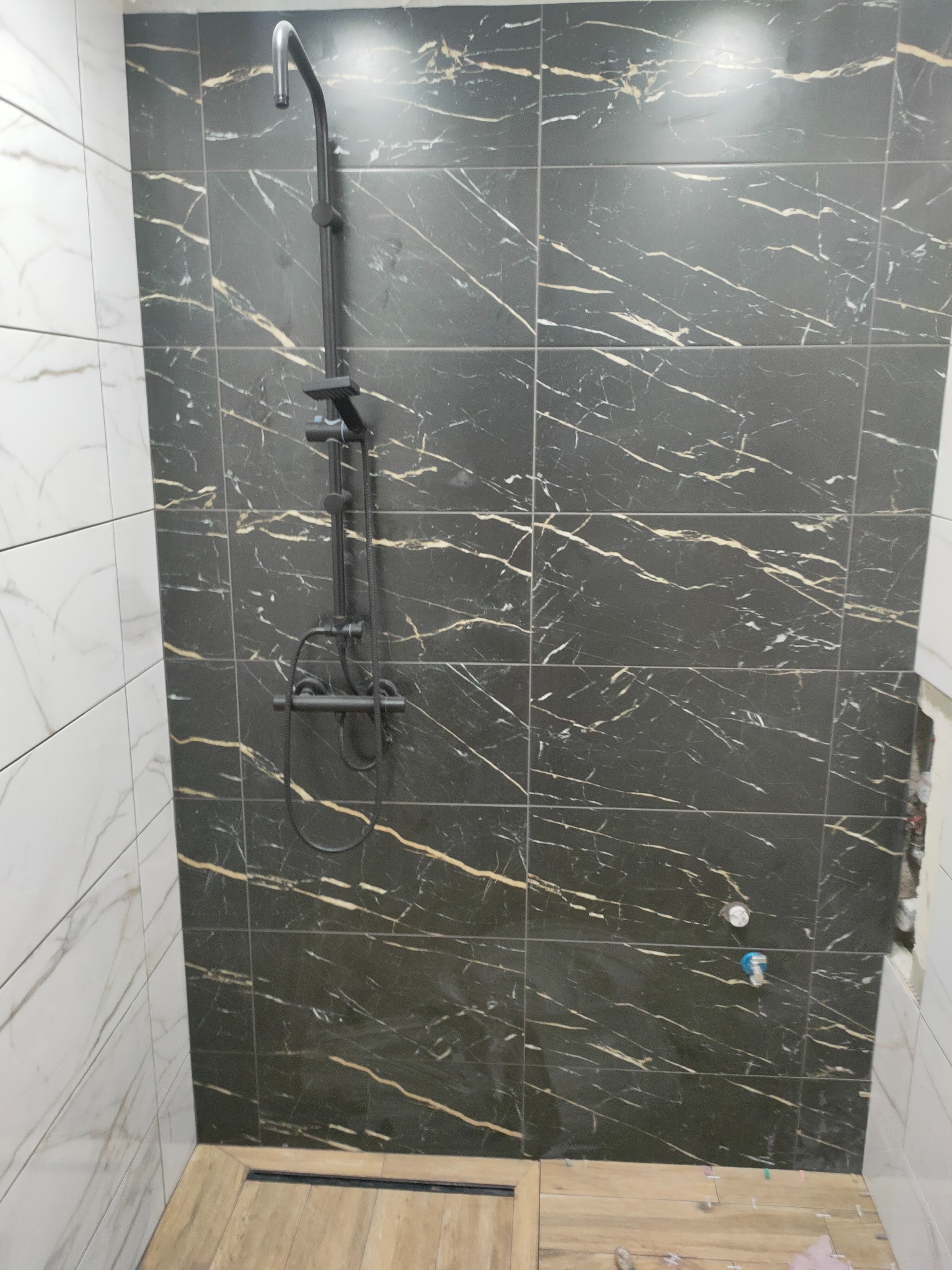 Kabina prysznicowa z czarnymi płytkami imitującymi marmur ze złotymi żyłami, zamontowana czarna armatura, widoczny fragment podłogi wyłożonej panelami imitującymi drewno, białe płytki na sąsiedniej...