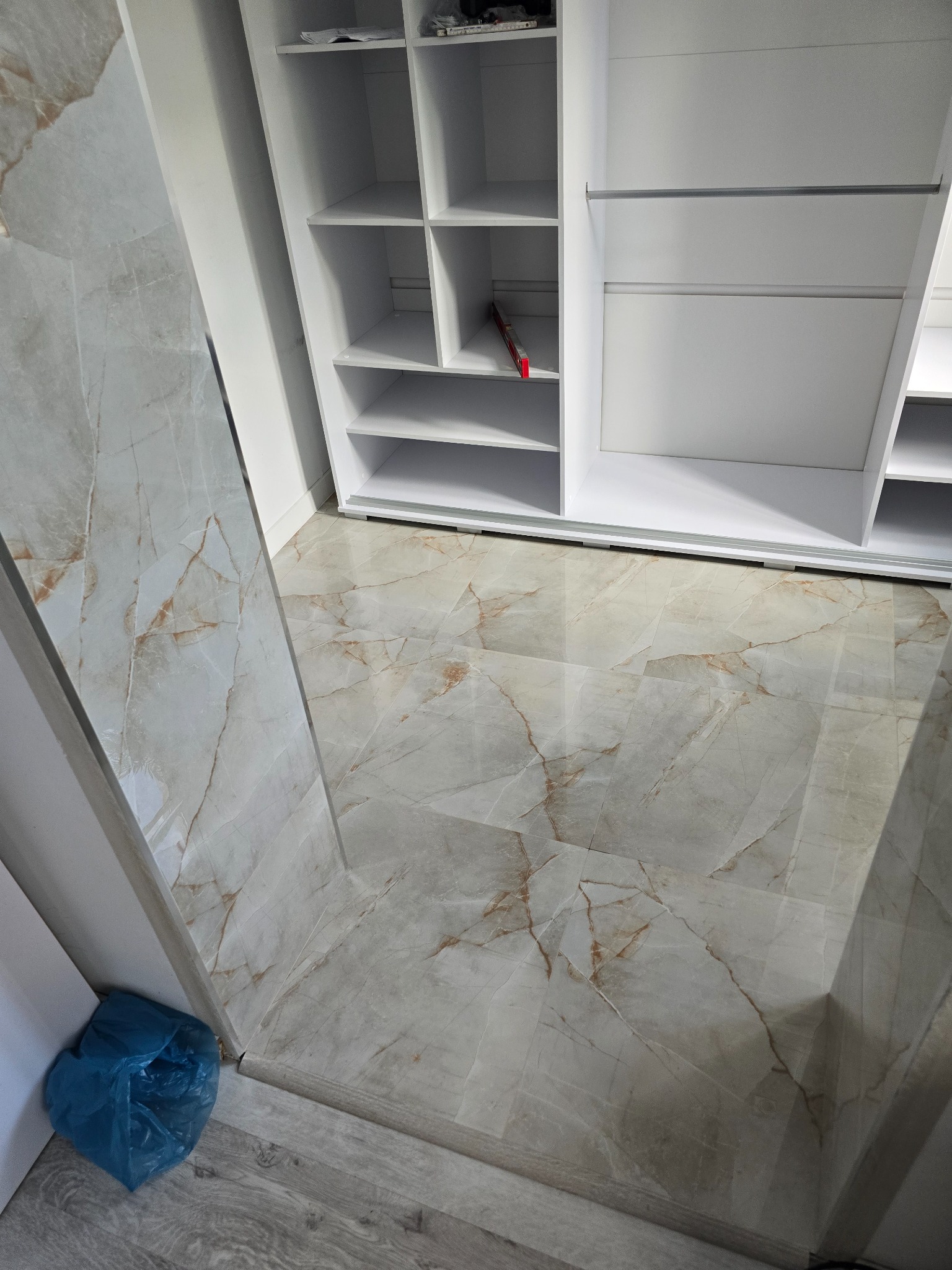 Ułożona podłoga z płytek imitujących marmur w odcieniach szarości i beżu, częściowo widoczna biała szafa wnękowa z półkami, w rogu niebieski worek na śmieci.