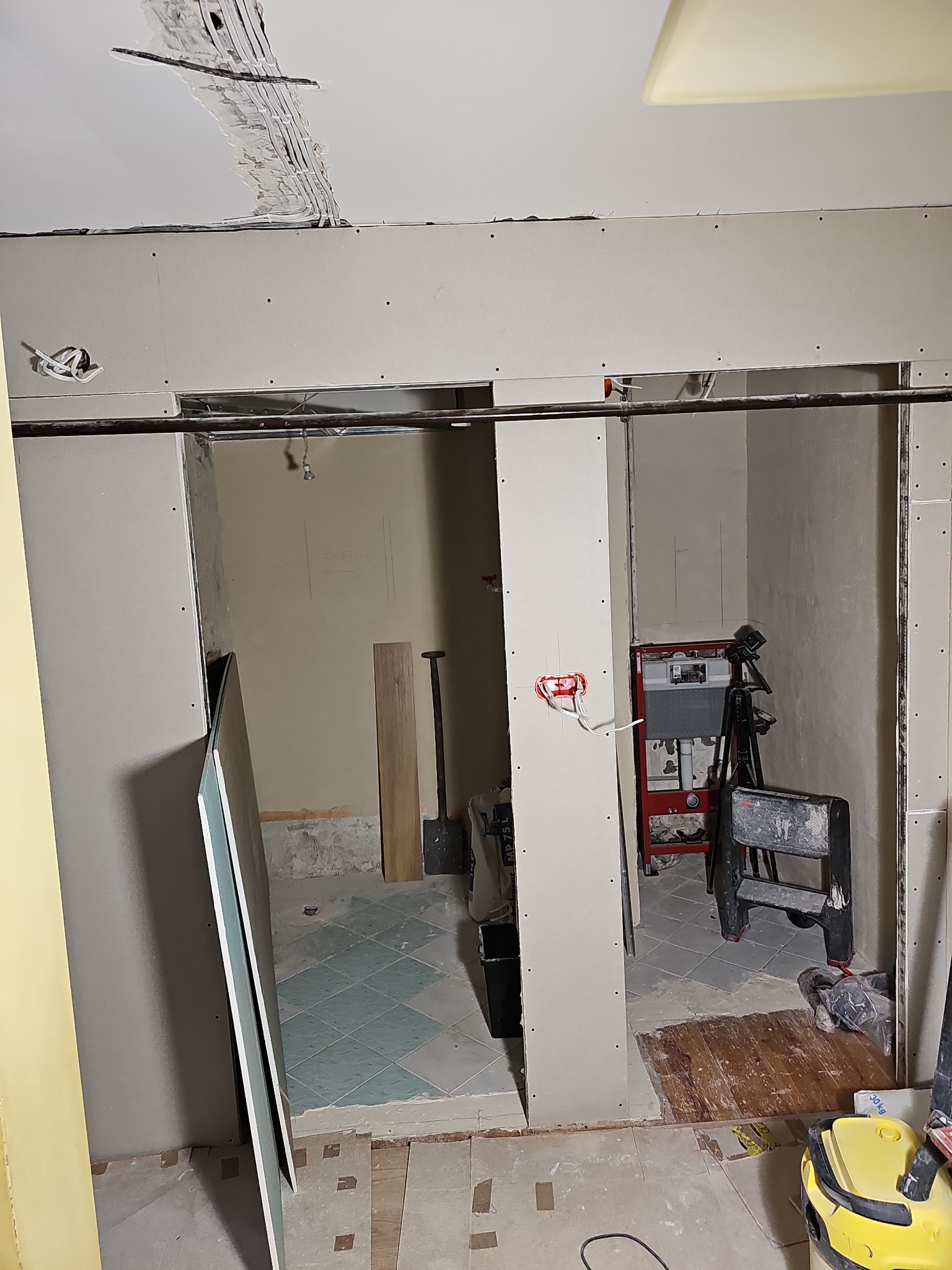 Łazienka w trakcie remontu: widoczne płyty gipsowo-kartonowe na ścianach, instalacja elektryczna, fragment starej podłogi z płytek i przygotowane podłoże pod nową posadzkę, drabina malarska...