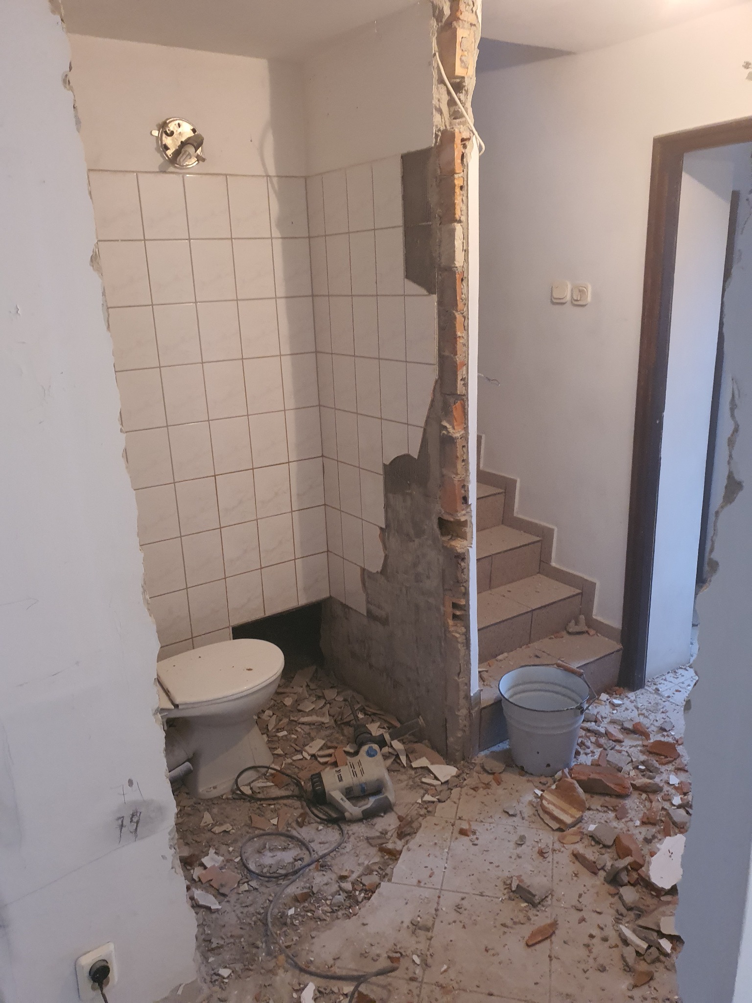 Demontaż ściany w łazience z widoczną toaletą, resztkami płytek, gruzem i narzędziami, prowadzący do schodów.