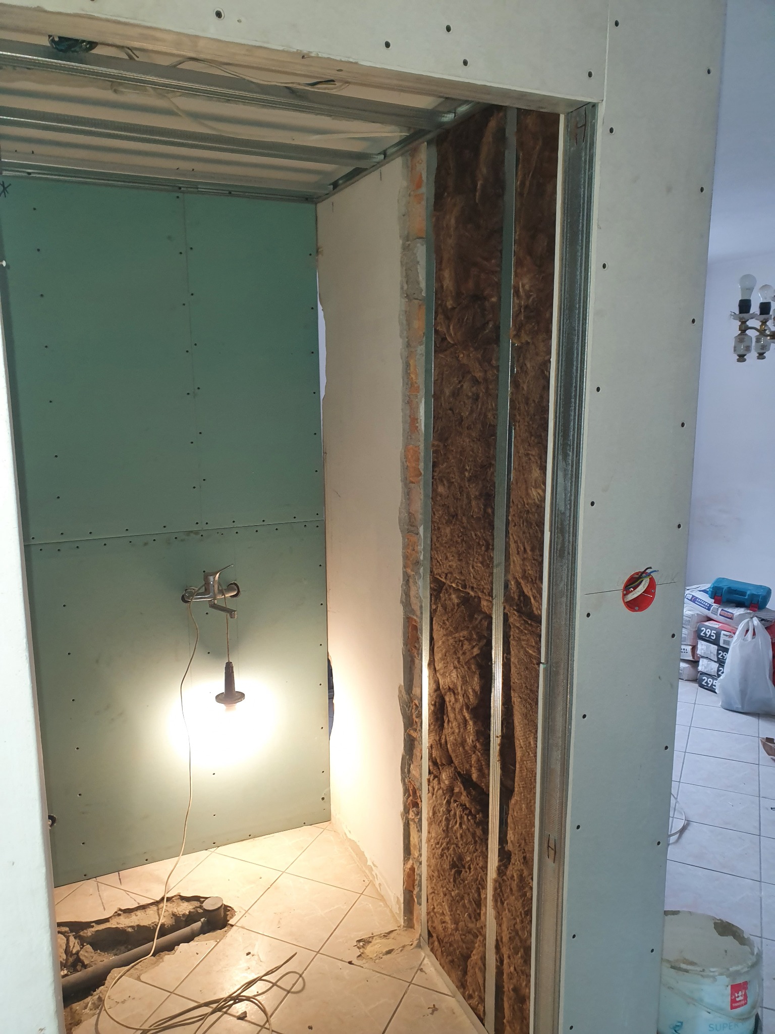 Wnętrze łazienki w trakcie prac w Piszu: widoczna konstrukcja metalowa, izolacja z wełny mineralnej w ścianie, płyty gipsowo-kartonowe i prowizoryczne oświetlenie halogenowe.