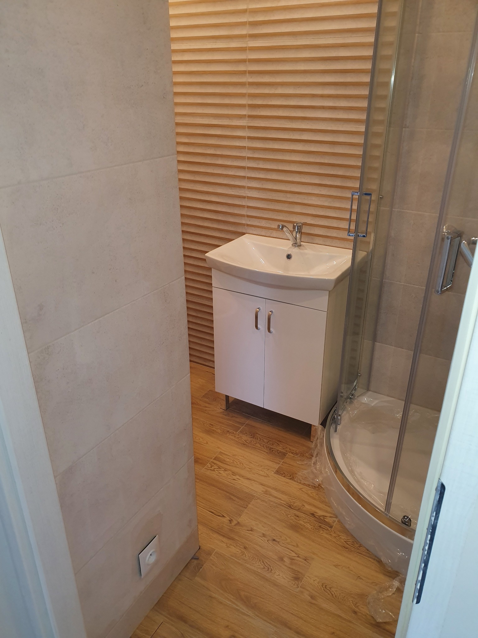 Nowoczesna łazienka z umywalką na szafce, kabiną prysznicową z zaokrąglonym narożnikiem i płytkami imitującymi drewno na ścianie oraz podłodze.