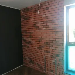 Fragment pokoju z dekoracyjną ścianą z cegieł, czarną ścianą obok, drewnianą podłogą i oknem z niebieską ramą, widoczne kable wystające z sufitu i rurki przy podłodze.