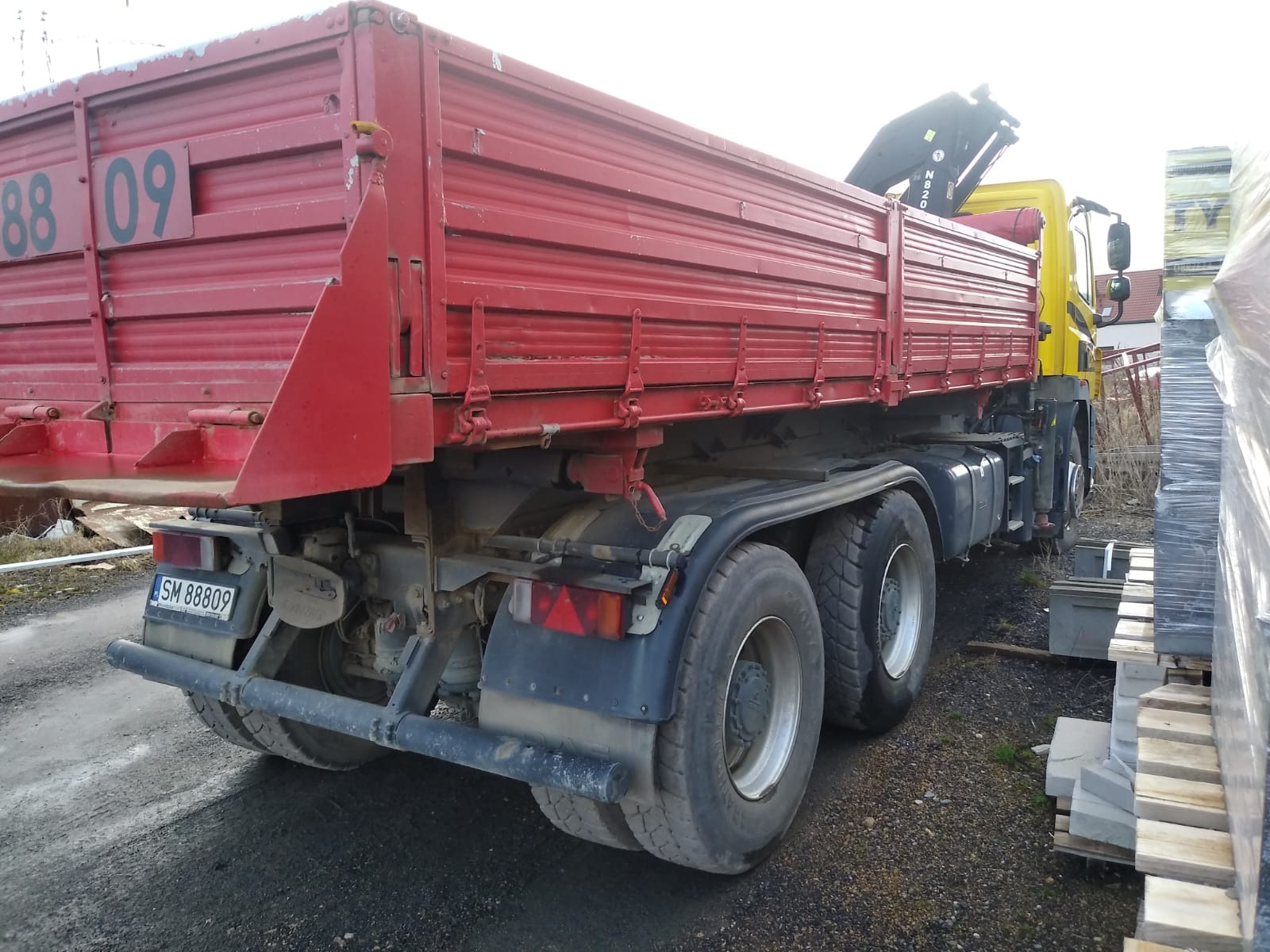 Czerwona wywrotka z żółtą kabiną i dźwigiem HDS, zaparkowana na placu z kostką brukową i paletami z materiałami budowlanymi.