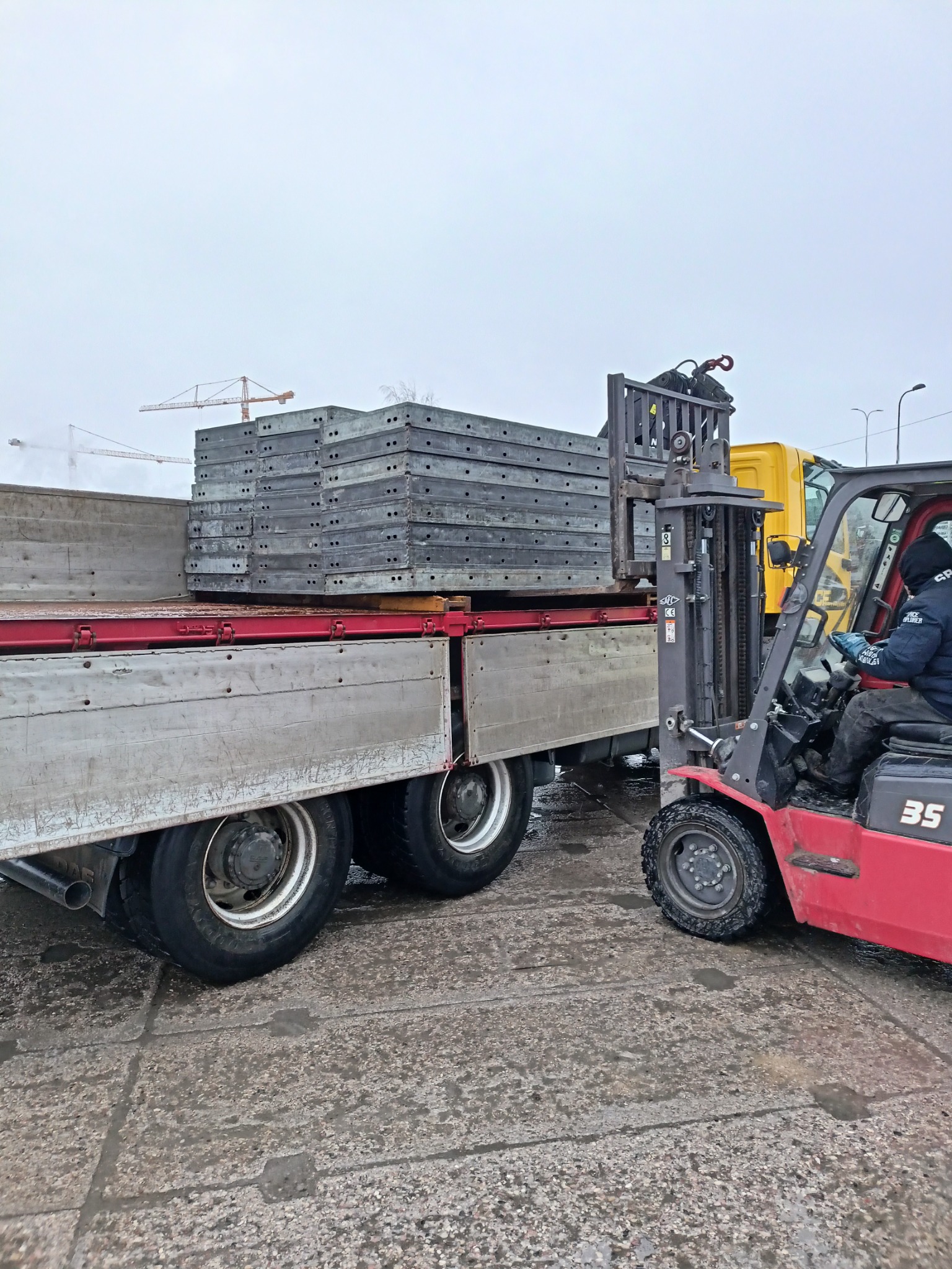 Czerwony wózek widłowy załadowuje paletę z szarymi, metalowymi płytami na skrzynię ciężarówki na tle budowy z dźwigami.