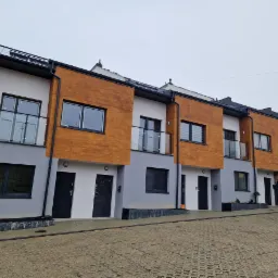 Szereg domów w zabudowie bliźniaczej z elementami elewacji w kolorze drewna i szarej tonacji, widok z brukowanej drogi dojazdowej.