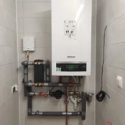 Nowoczesny, biały piec gazowy marki Buderus zamontowany na ścianie wyłożonej jasnymi płytkami, z widocznymi rurami i elementami instalacji grzewczej, w tym zaworami i pompą obiegową...