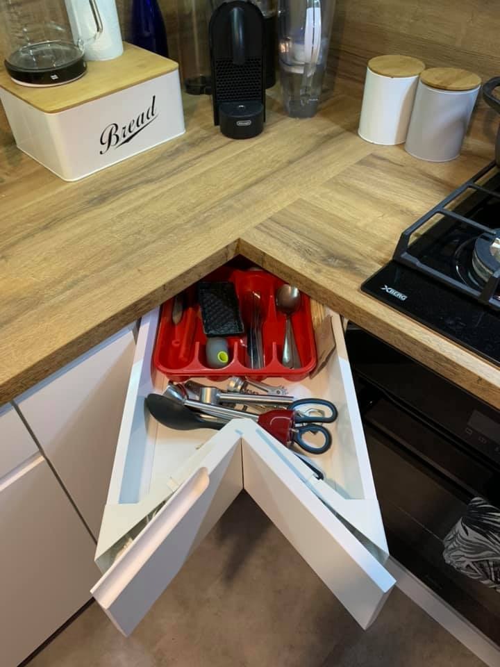 Narożna szuflada z przegródkami na sztućce i akcesoria kuchenne, drewniany blat w jodełkę, sprzęt AGD w tle.