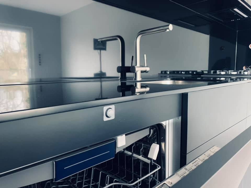 Nowoczesna kuchnia z otwartą zmywarką, czarnym blatem i minimalistyczną armaturą, odbicie w szklanym panelu ściennym.