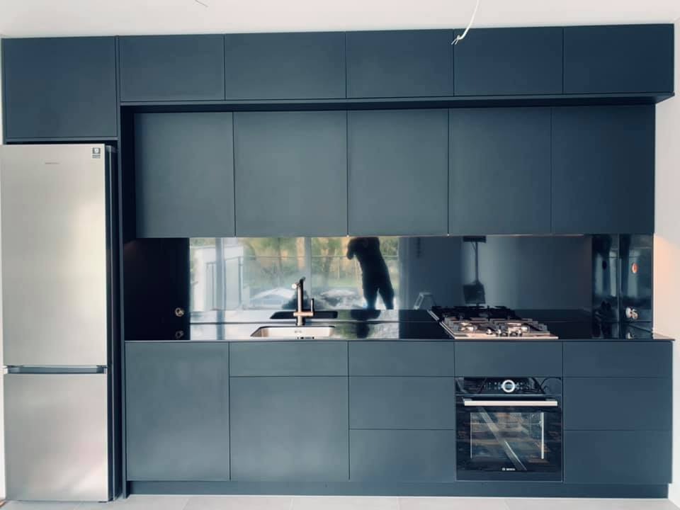 Nowoczesna kuchnia w odcieniach szarości z zabudową meblową pod sufit, czarnym blatem i lustrzanym panelem nad zlewem, z widocznym odbiciem osoby fotografującej.