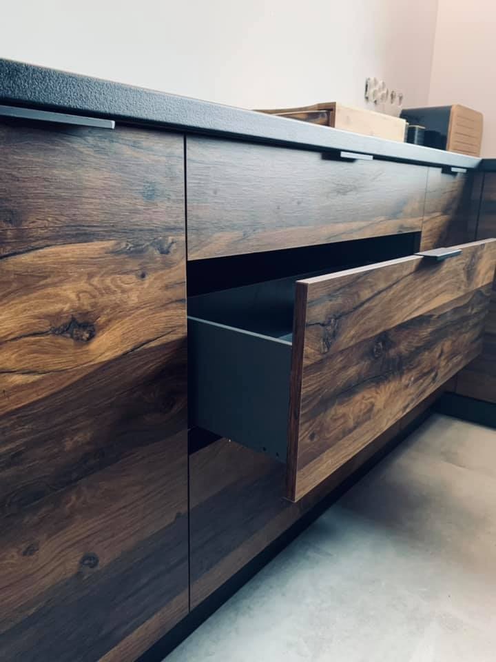 Szczegółowe ujęcie szuflady z ciemnego drewna o wyraźnym usłojeniu, wysuniętej z zabudowy kuchennej z czarnym blatem, minimalistyczne uchwyty.