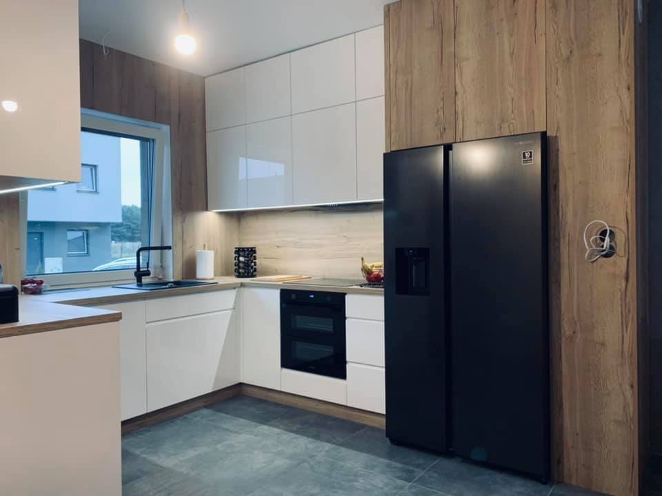 Nowoczesna kuchnia z białymi szafkami, drewnianymi panelami ściennymi i czarną lodówką side-by-side. Oświetlenie LED podszafkowe. Widok na budynek za oknem.