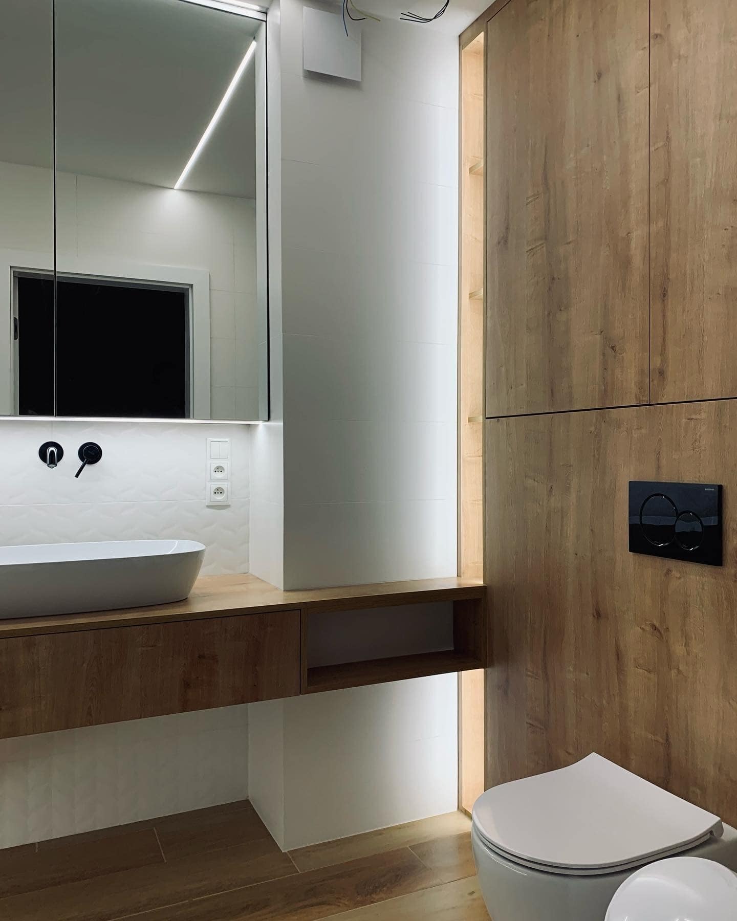Nowoczesna łazienka z drewnianymi szafkami, umywalką nablatową, podświetlanym lustrem i wiszącą toaletą z czarnym panelem