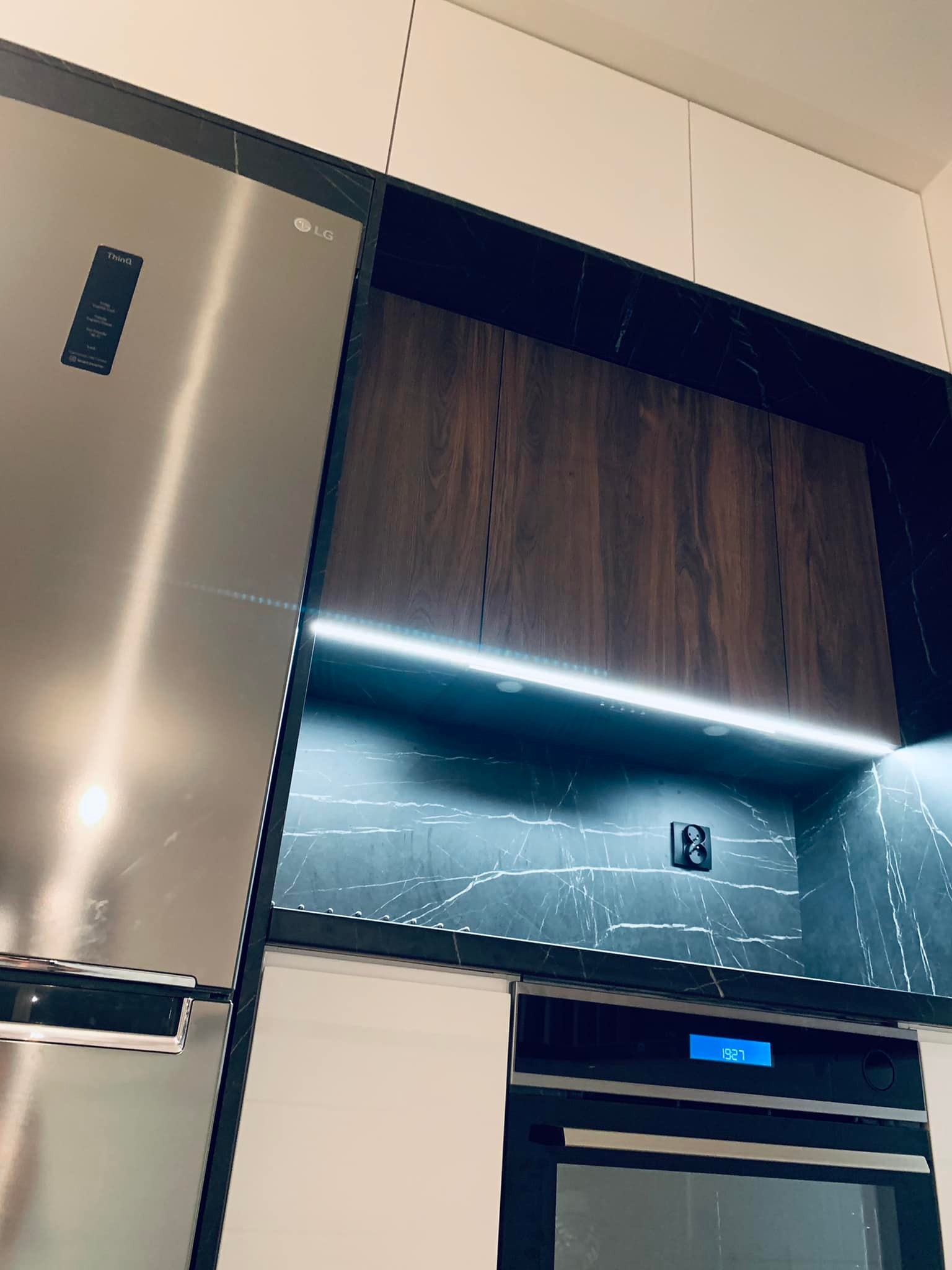 Nowoczesna zabudowa kuchenna z frontami w kolorze drewna i czarnym marmurem, podświetlenie LED nad blatem, obok lodówka LG w stali nierdzewnej, widoczny piekarnik z wyświetlaczem.