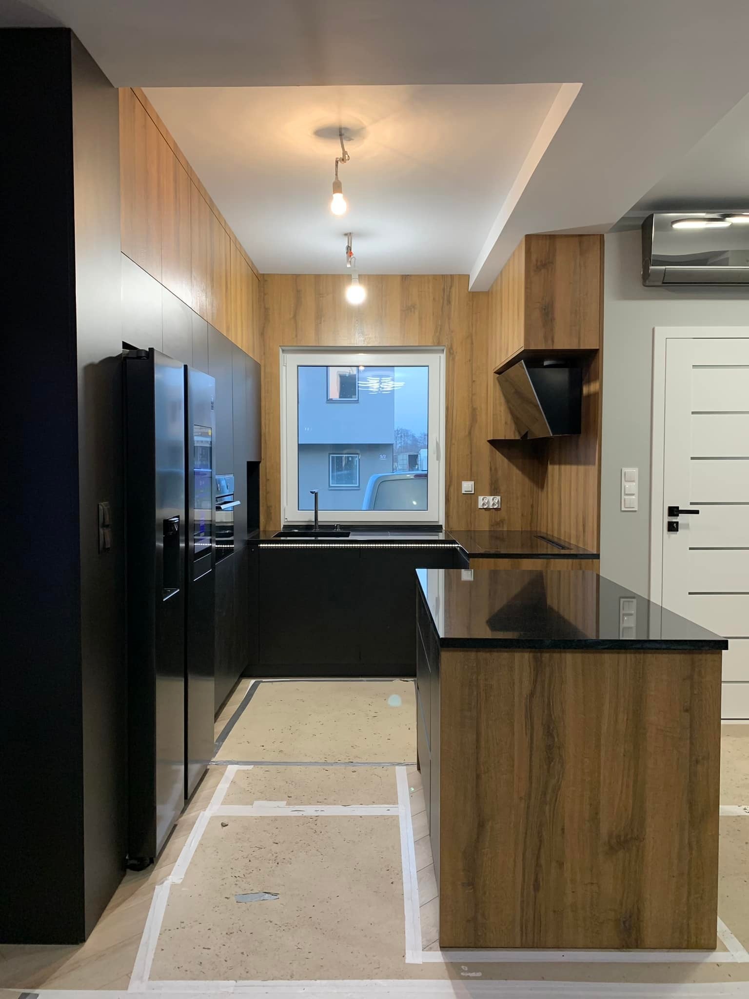 Nowoczesna kuchnia w trakcie montażu: czarne fronty szafek, drewnopodobne wykończenie ścian i wyspy, czarny blat, widoczne taśmy malarskie na podłodze.