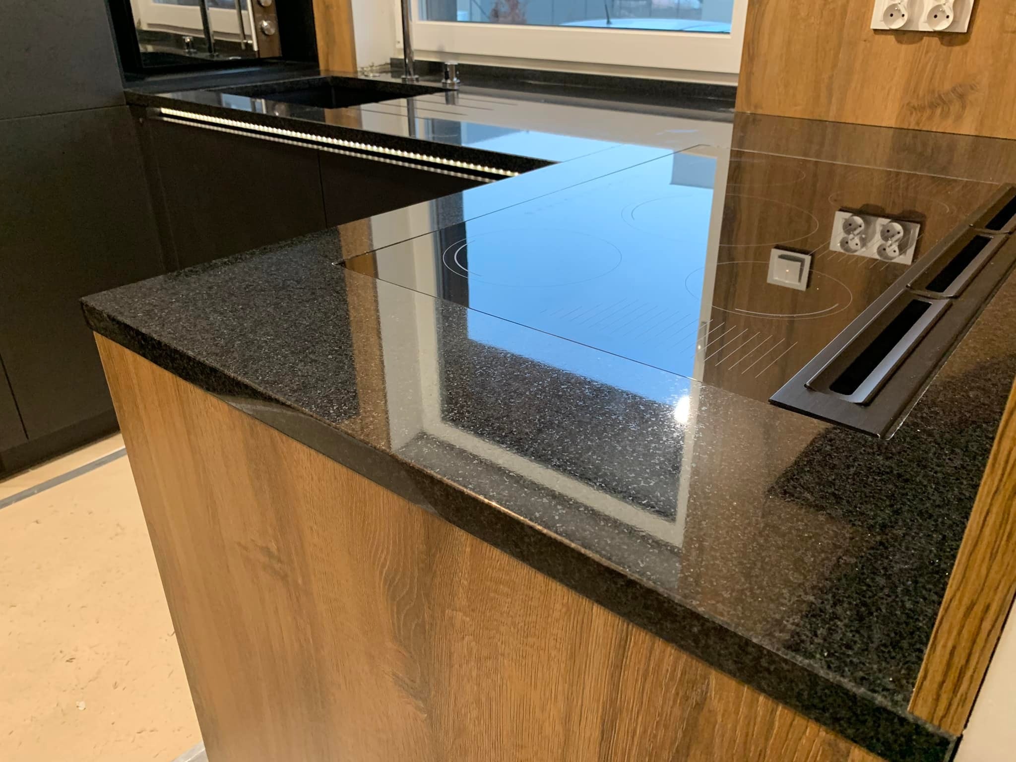 Czarny, błyszczący blat z kamienia z wbudowaną płytą indukcyjną i pochłaniaczem pary, fronty szafek w kolorze drewna, gniazdka elektryczne na ścianie.
