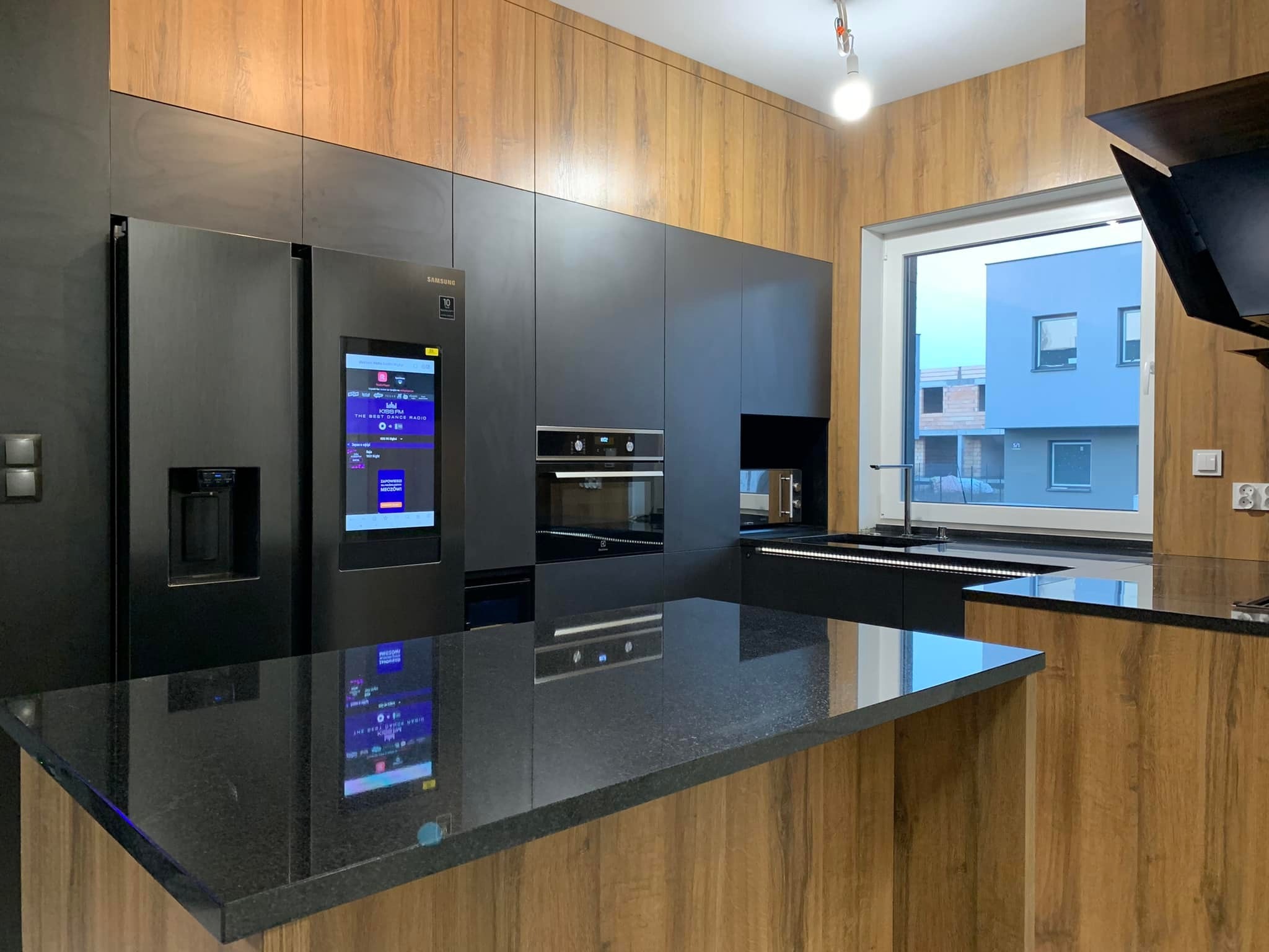 Nowoczesna kuchnia z czarnymi frontami szafek i drewnopodobnymi panelami ściennymi, wyspa z czarnym blatem, lodówka Samsung z wyświetlaczem, widok na budynek za oknem.