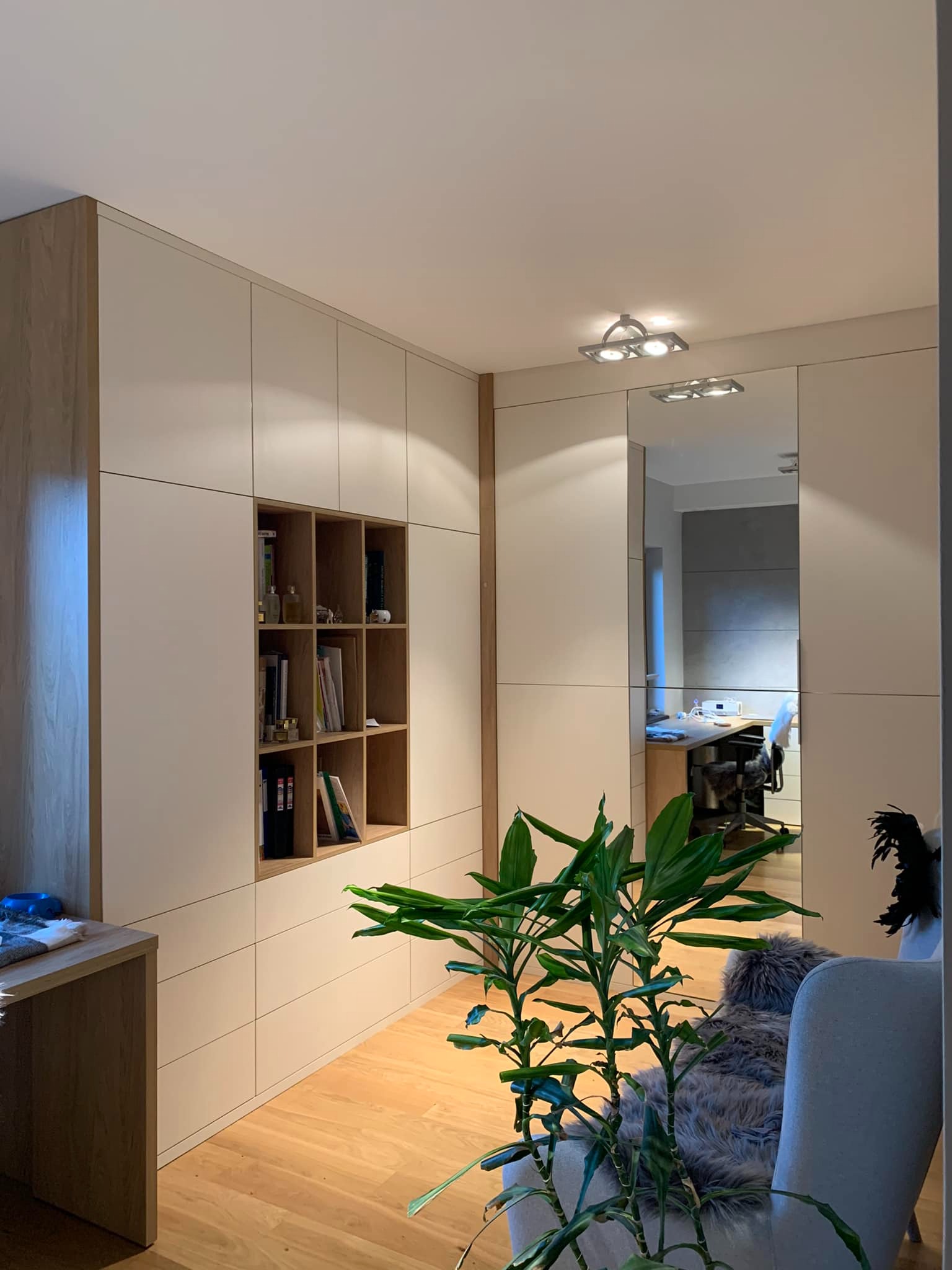 Białe szafy wnękowe z drewnianymi akcentami i otwartą półką na książki, widok z perspektywy pokoju z rośliną doniczkową i odbiciem biurka w lustrzanych drzwiach.
