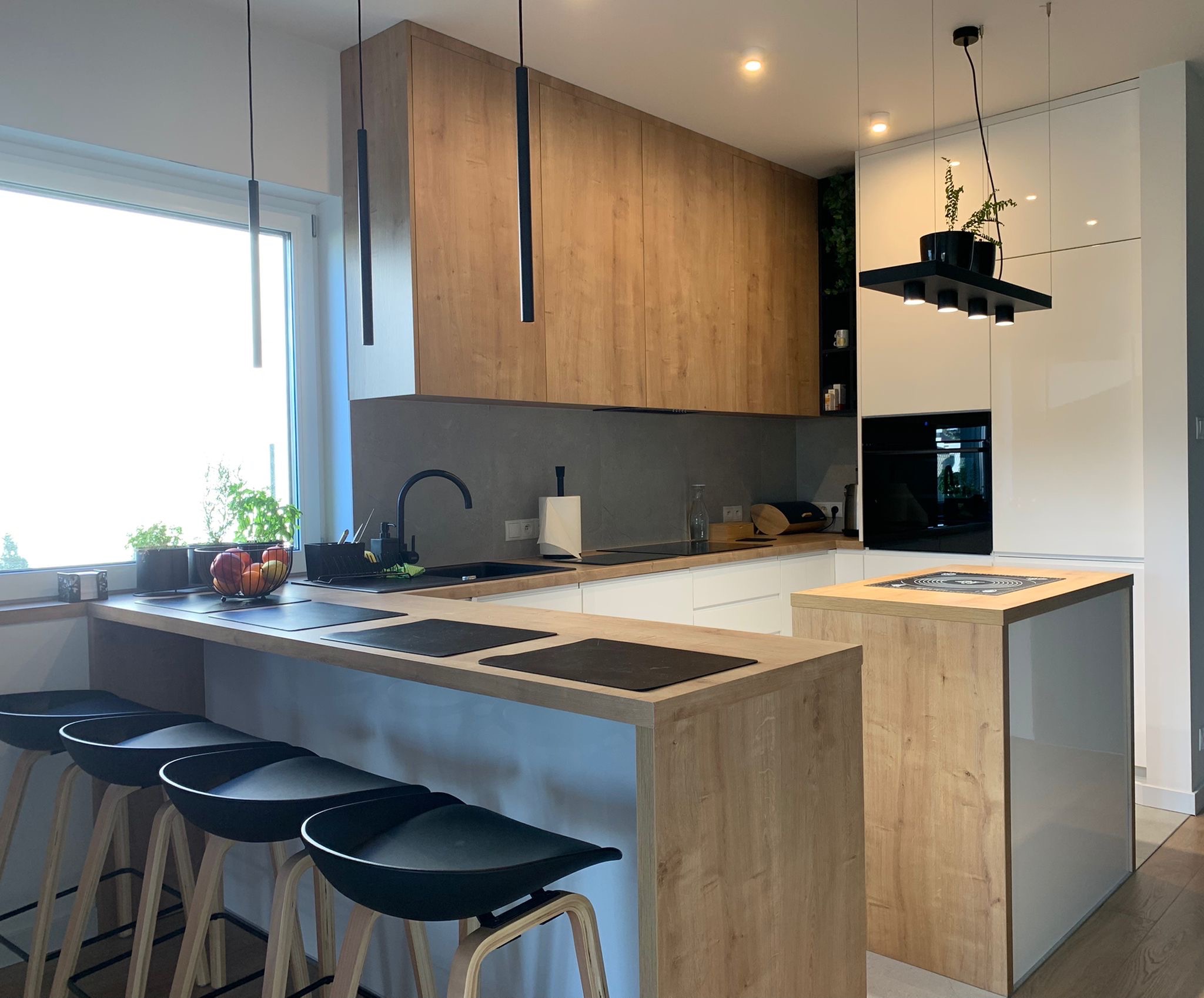 Nowoczesna kuchnia z drewnianymi frontami szafek górnych i wyspą, białe szafki dolne, czarne hokery i lampy wiszące, rośliny w oknie i na lampie.