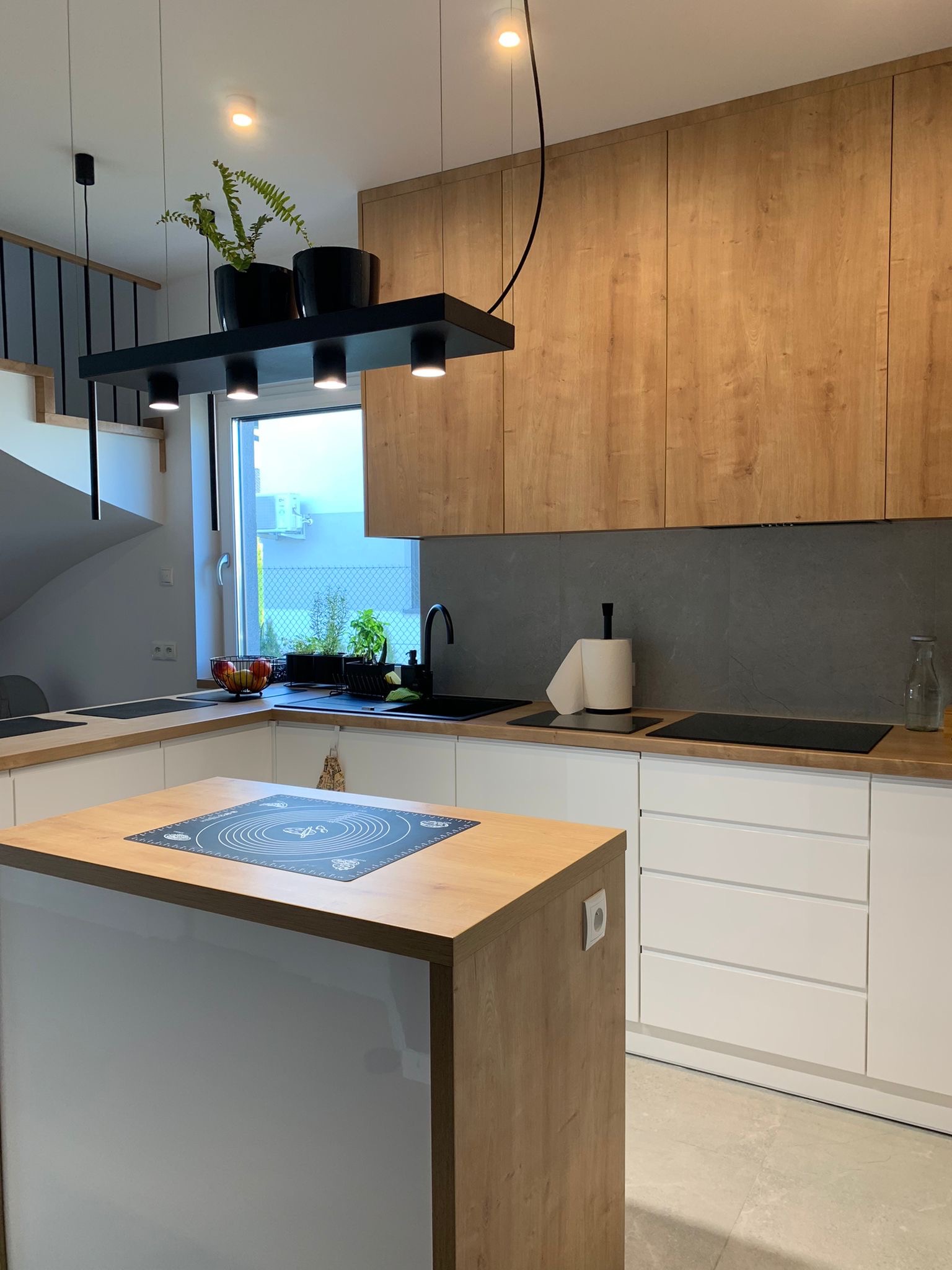 Nowoczesna kuchnia z wyspą, drewniane fronty szafek górnych i blatu wyspy, białe fronty dolne, czarny zlew i bateria, czarne lampy wiszące nad wyspą z doniczkami, szara ściana nad blatem.
