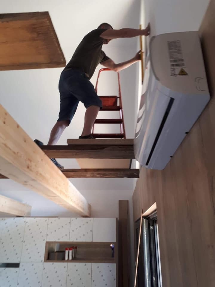 Mężczyzna instaluje klimatyzator na ścianie nad drewnianą antresolą, używając poziomicy, z widoczną drabiną i częścią zabudowy kuchennej w tle.
