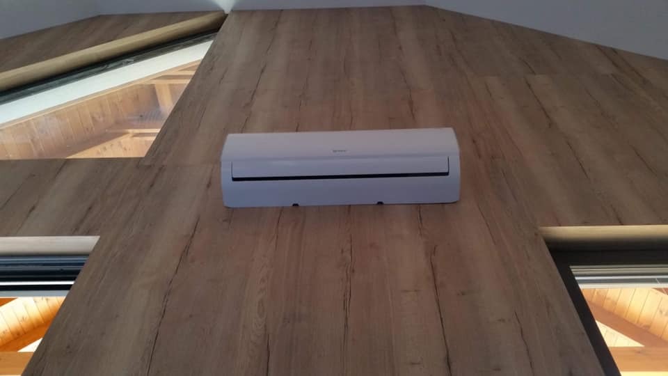 Biała klimatyzacja zamontowana na skośnej ścianie obłożonej panelami imitującymi drewno, z widocznym oknem dachowym w tle.
