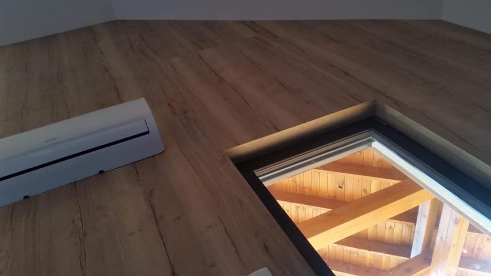 Drewniana podłoga na poddaszu ze skośnym oknem dachowym, przez które widać drewnianą konstrukcję dachu, oraz zamontowany klimatyzator.