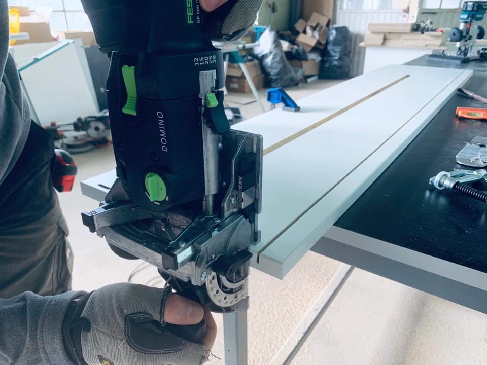 Frezarka lamelowa Festool Domino DF 500 w trakcie pracy nad płytą meblową z frezem, widoczne rękawice robocze i akcesoria stolarskie na blacie.