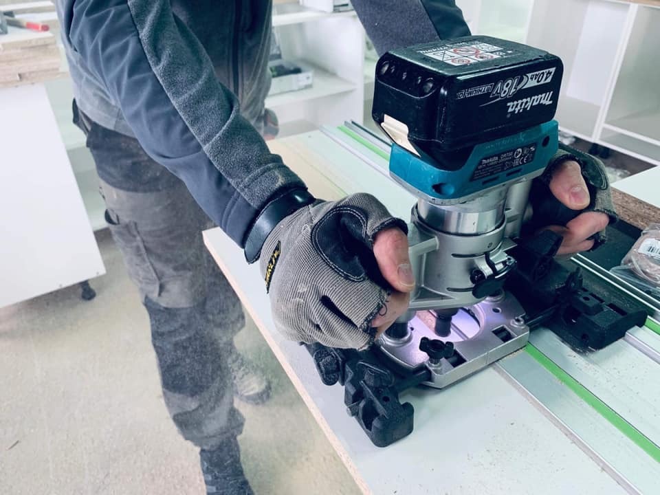 Stolarz frezuje krawędź płyty meblowej za pomocą frezarki Makita z akumulatorem 18V, używając szyny prowadzącej dla precyzji, w tle widoczne elementy białych mebli.