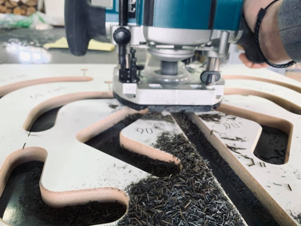 Frezarka górnowrzecionowa w trakcie wycinania kształtu w białej płycie MDF, widoczne wióry i fragment dłoni operatora w rękawicy.