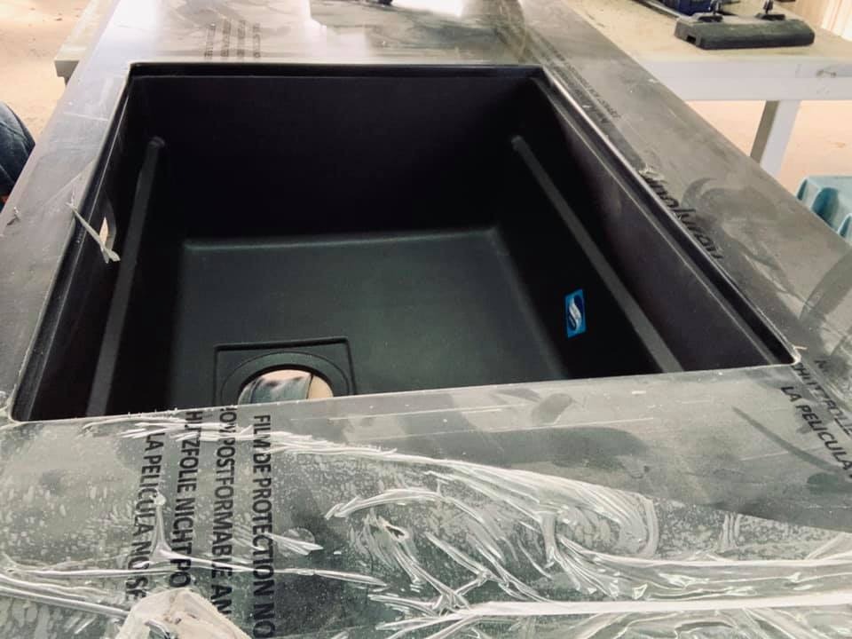 Czarny zlew wpuszczony w blat, zabezpieczony folią ochronną z napisami, widoczne narzędzia w tle.