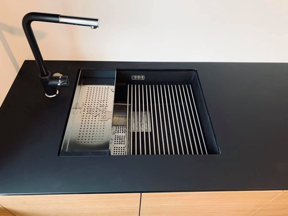 Czarny, minimalistyczny blat kuchenny z wbudowanym zlewem z akcesoriami: ociekaczem, rusztem i czarną baterią. Widoczne drewniane fronty szafek pod blatem.