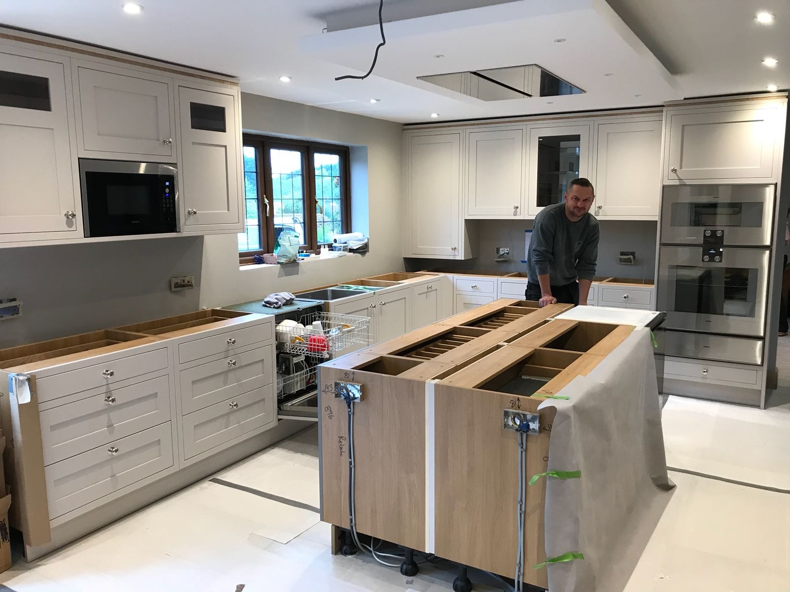 Montaż jasnej kuchni: wyspa kuchenna w trakcie instalacji, widoczne okablowanie, szafki bez blatów, mężczyzna w tle kontroluje postęp prac.