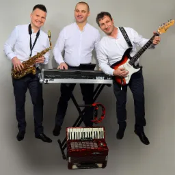 Trzech uśmiechniętych muzyków w białych koszulach i granatowych spodniach pozuje z instrumentami: saksofonem, keyboardem Korg Pa4X i gitarą elektryczną, a pod keyboardem leży czerwony akordeon...
