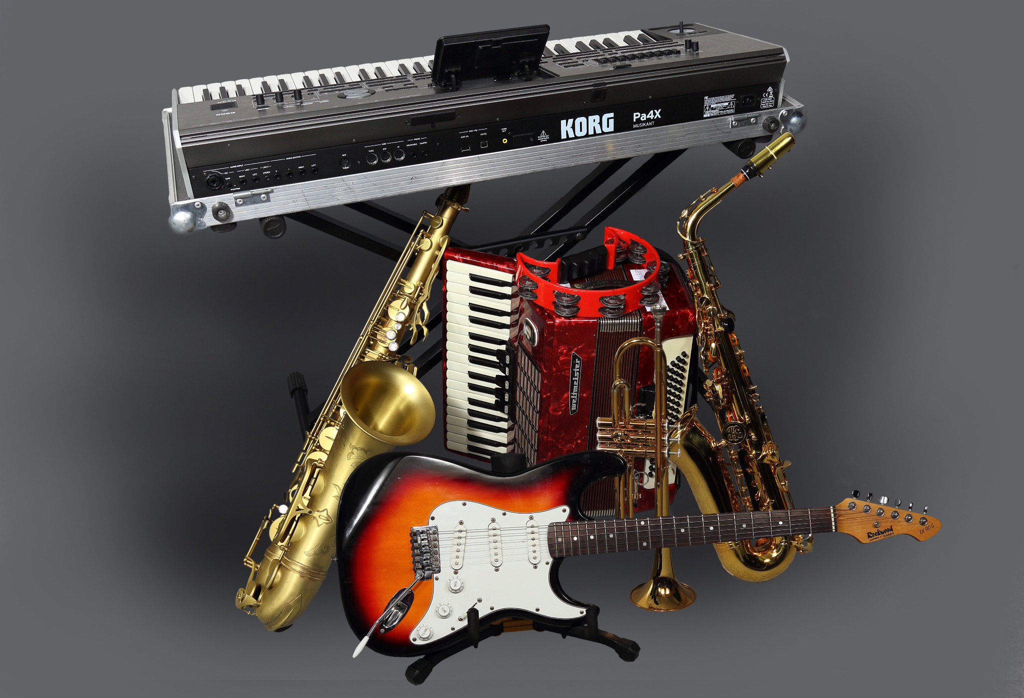 Różnorodność instrumentów muzycznych: keyboard Korg, saksofony, akordeon, trąbka i gitara elektryczna Rockwood, ustawione na tle szarego studia.